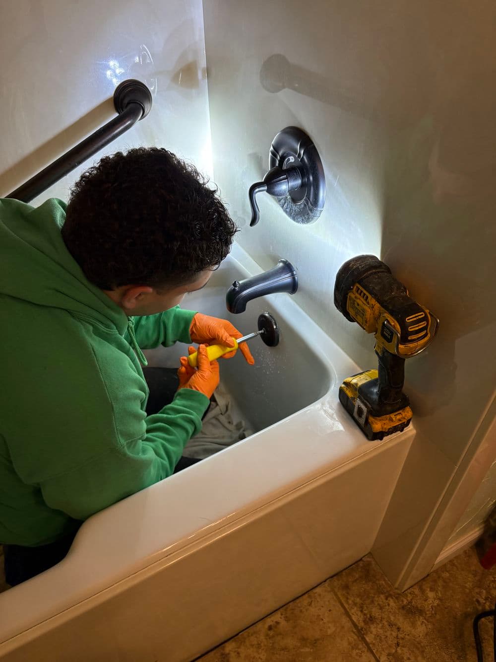 Person in green hoodie performing plumbing repairs in a bathtub with tools.